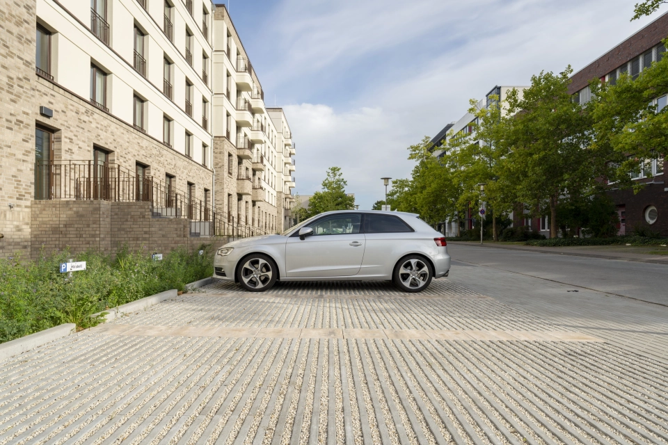 Parkplatz vor einem modernen Gebäude mit beiger Fassade im oberen Bereich und braunen Wandfiesen im unteren Bereich. Die Parkfläche ist mit grauen Rasenfugensteinen und beigen Betonsteinen, zur Abgrenzung der einzelnen Parkplätze, gepflastert. Ein silbernes Auto parkt auf einem Stellplatz. Drumherum sind grüne Bäume, Sträucher und Gräser zu finde.