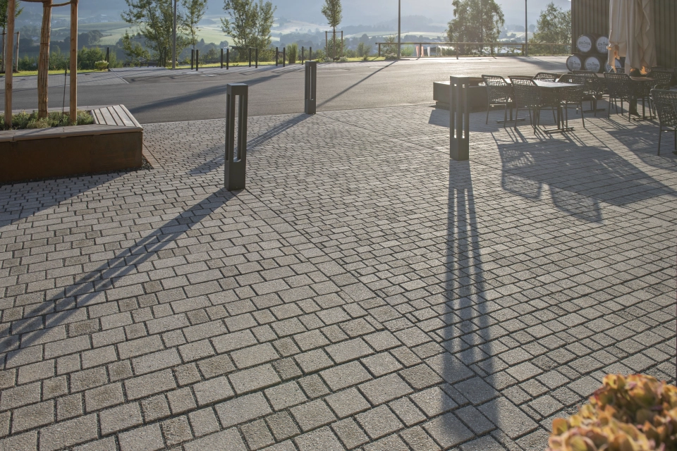 Nahaufnahme von beigen Pflastersteinen. Im Hintergrund sind Tische, Stühle, Lichtstehlen und eine geteerte Straße.