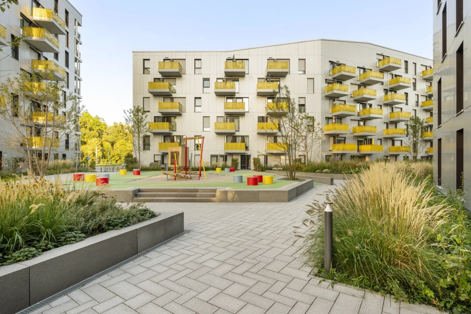 Innenhof einer Wohnanlage mit mehrgeschossigen Gebäuden. Die Wohngebäude haben gelbe Balkonbrüstungen und eine helle Fassade. Im Innenhof befinden sich einige begrünte Beete und auf einer erhöhten Fläche ist ein bunter Kinderspielplatz. Die Fläche ist mit basatlt- und granitfarbenem GDM.SCENIC individual gepflastert.