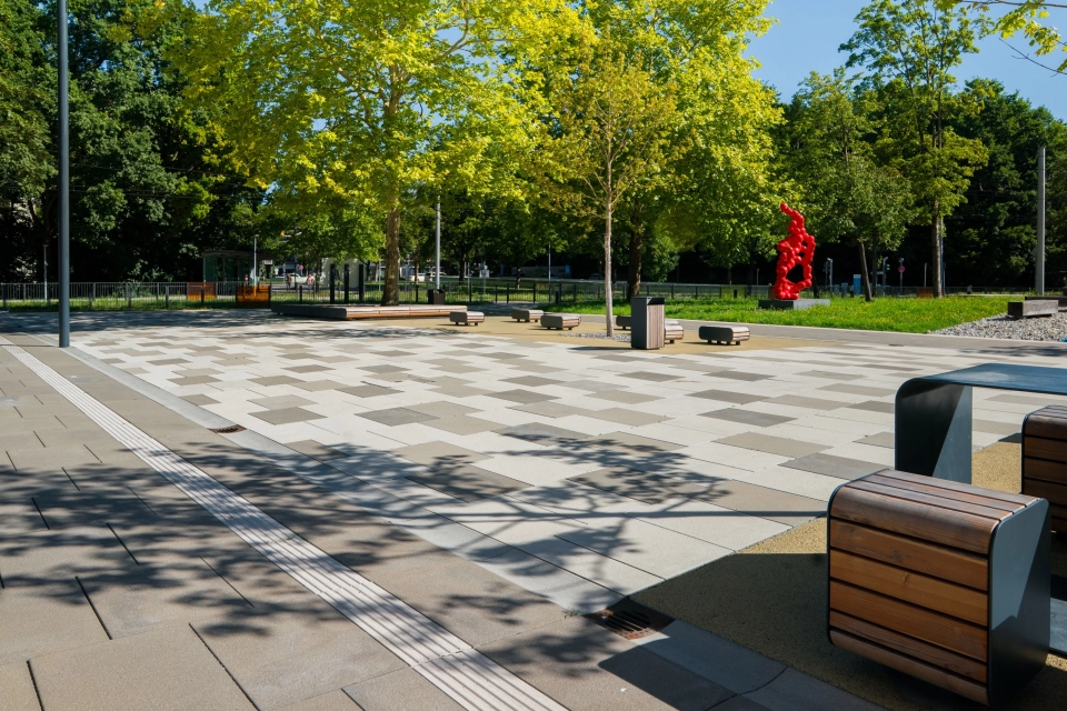 Offener Platz auf einem Universitätscampus mit Bäumen, Sitzbänken und einer roten Skulptur im Hintergrund, wobei der Platz mit GDM.SCENIC stone in drei unterschiedlichen Farbabstufungen gepflastert ist.
