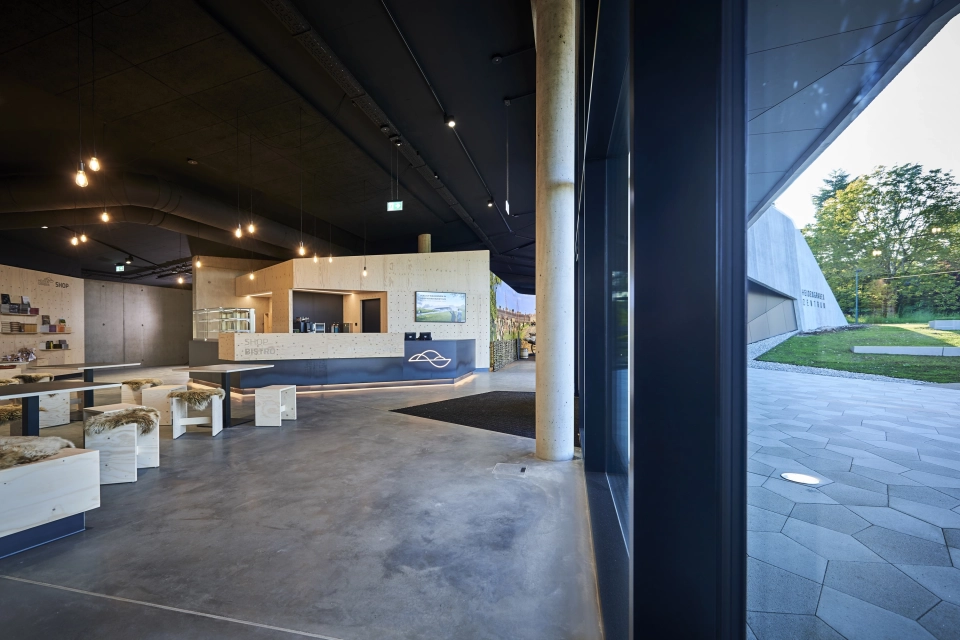 Foyer eines Gebäudes mit hölzernem Empfang und Sitzmöglichkeiten. Beim Blick durch die Fenster sind draußen fünfeckige Betonsteine zu erkennen.