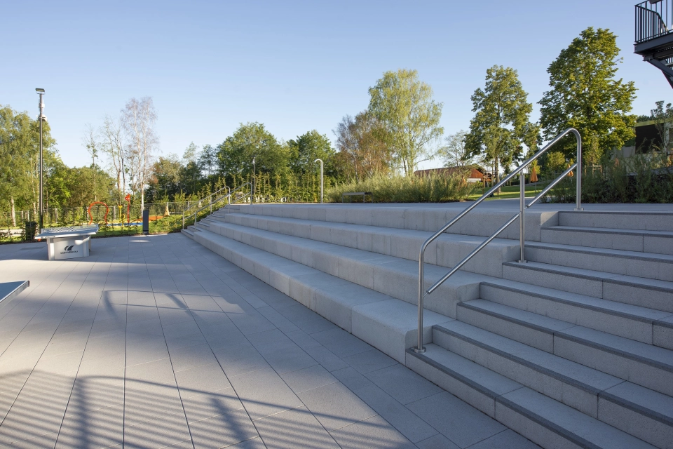 Breite, mehrstufige Treppenanlage aus GDM.SCENIC step führt zu einer offenen Terrasse, auf der Betonsteine verlegt sind; umgeben von Grünflächen und Bäumen unter klarem Himmel.
