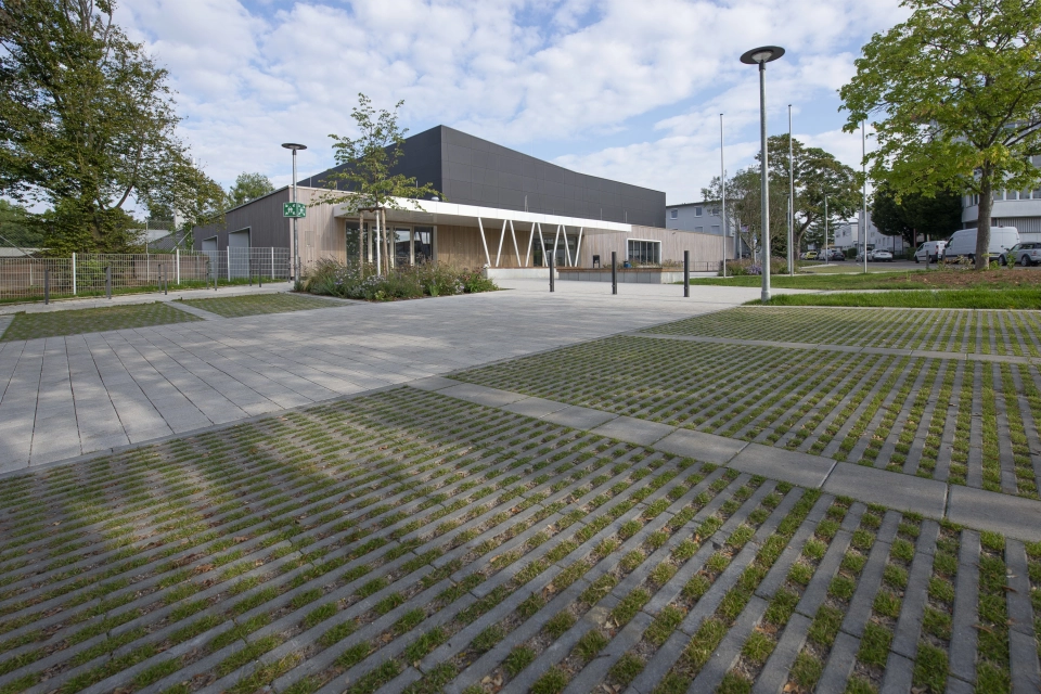 Im Vordergrund ist ein Parkplatz aus begrünten Rasenfugensteinen. Dahinter ist ein Weg aus Betonsteinen und eine moderne Sporthalle mit anthrazitfarbener Fassade und Holzverkleidung. Drumherum sind grüne Bäume und Sträucher zu finden.