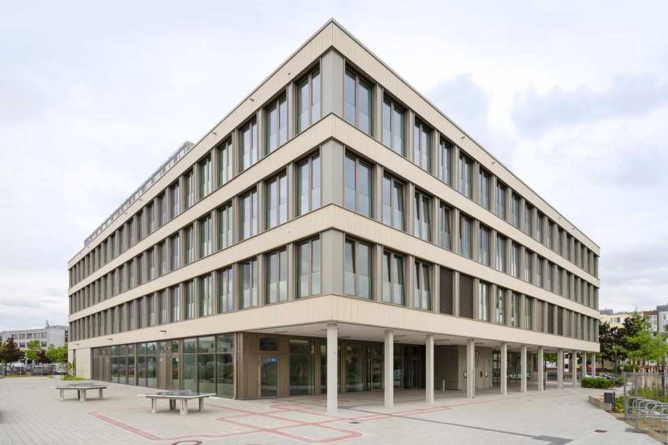 Offener Schulhof mit Tischtennisplatten und einer grau-beigen gepflasterten Fläche. Im Fokus steht ein modernes Schulgebäude mit großen Fenstern.