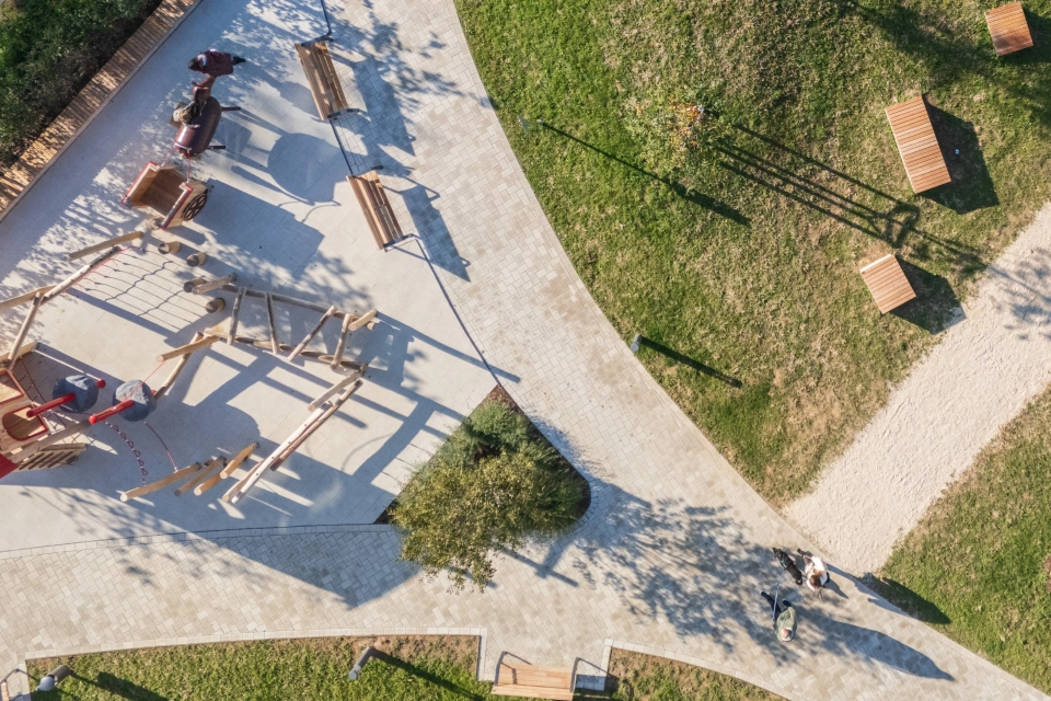 Luftaufnahme eines modernen Spielplatzbereichs mit hellen Wegen aus beigem GDM.KLIMASTEIN, umgeben von Grünflächen, Sitzbänken und hölzernen Spielgeräten.