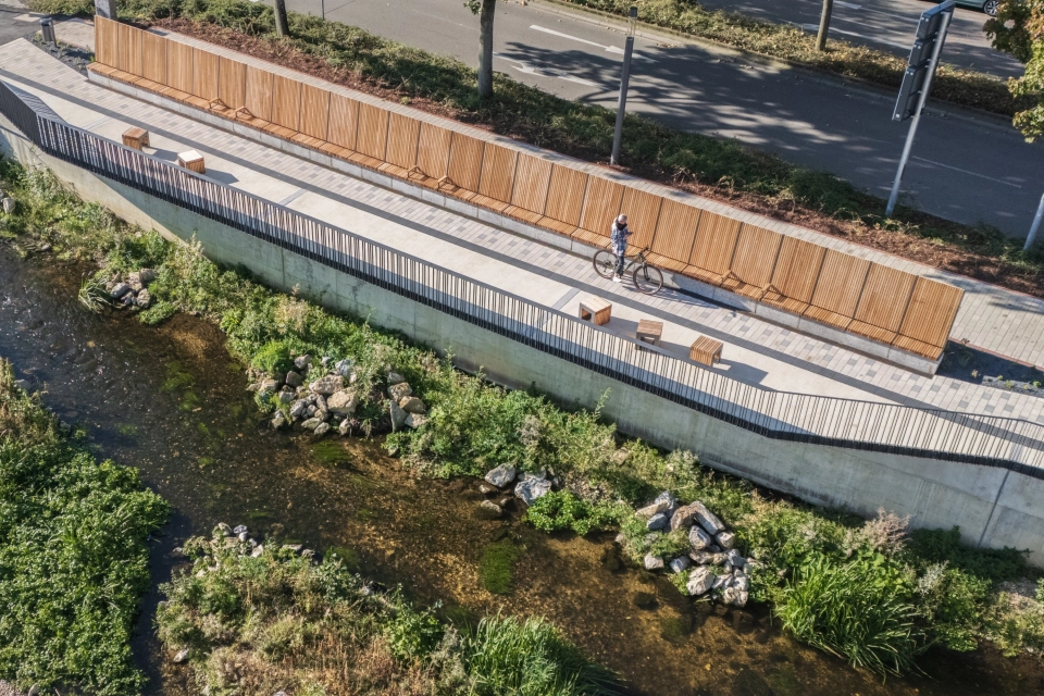 Luftaufnahme eines Uferwegs entlang eines flachen Bachlaufs, gesäumt von Holzsitzbänken und einer langen Holzverkleidung. Der Weg ist mit hellem Betonstein gepflastert, während oberhalb eine Straße mit Bäumen verläuft.