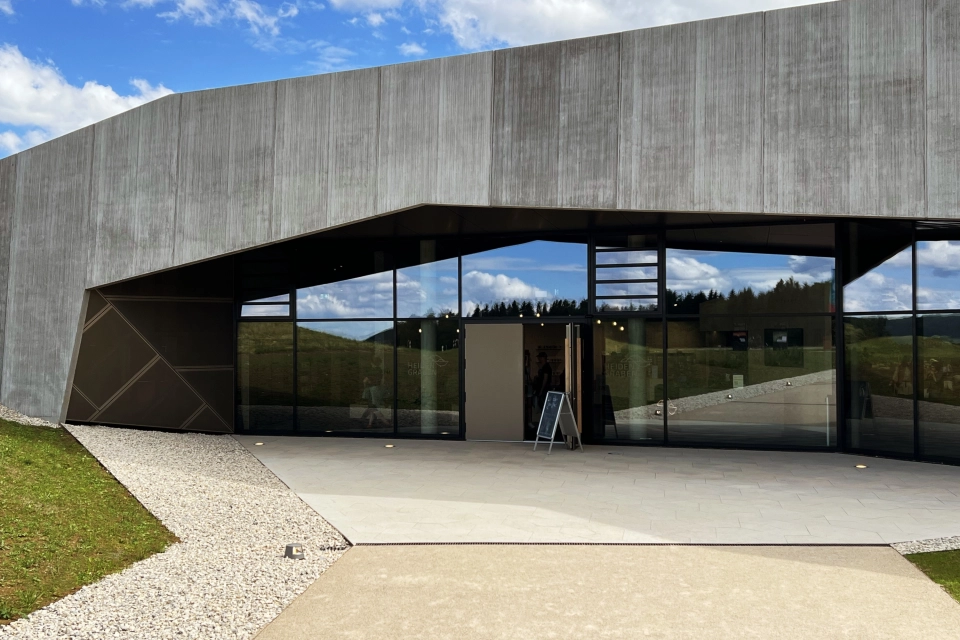 Nahaufnahme einer Gebäudefassade mit Holzoptik und Fensterfront. Davor sind fünfeckige, kalksteinfarbene Betonsteine verlegt.