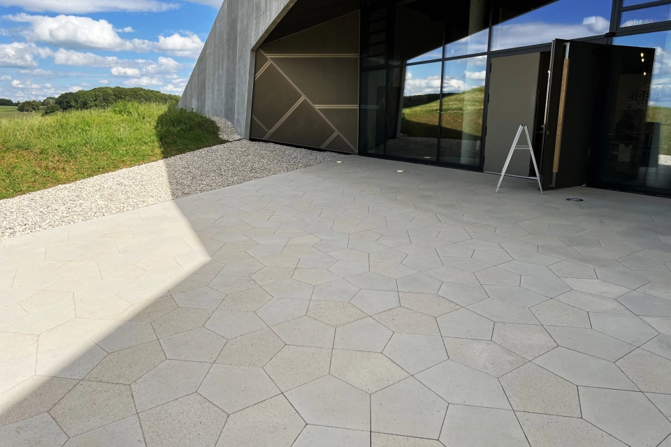 Nahaufnahme von kalksteinfarbenen, fünfeckigen Betonsteinen. Im Hintergrund ist eine Grünfläche und ein modernes Gebäude mit großen Fenstern.