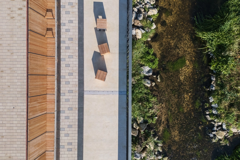 Draufsicht auf einen Uferweg mit mehreren Holzsitzbänken, angrenzend an einen naturbelassenen Bachlauf mit Steinen und dichtem Uferbewuchs. Der Weg ist mit beigem GDM.KLIMASTEIN gepflastert.