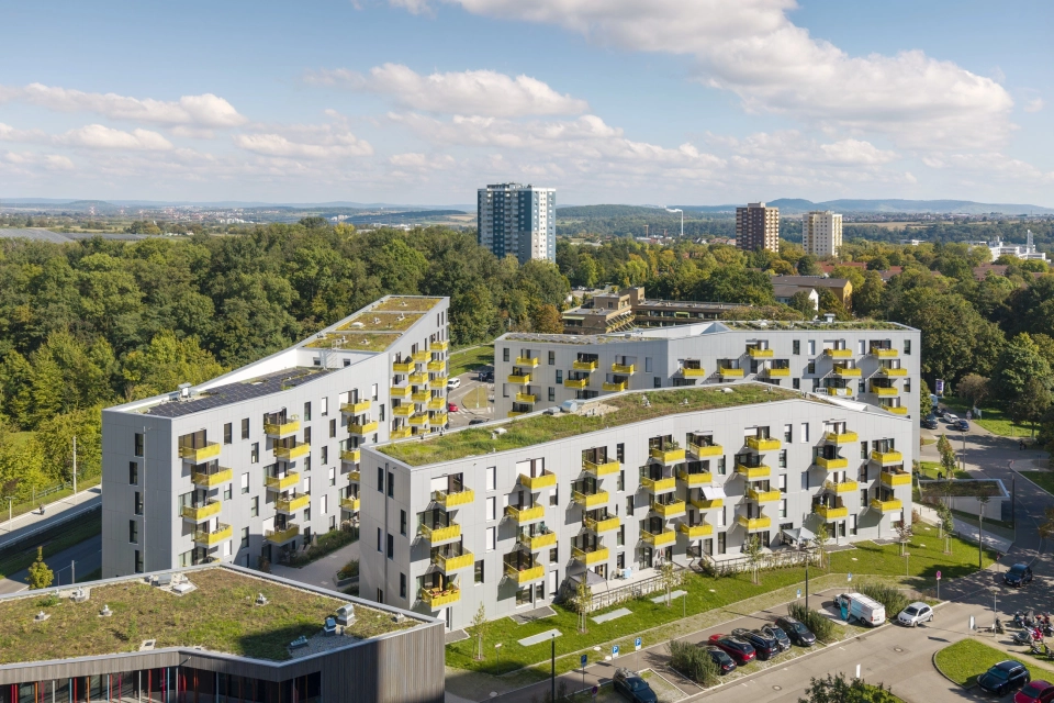 Drei moderne, mehrgeschossige Wohnhäuser mit heller Fassade, gelben Balkonbrüstungen und begrünten Dächern. Die Gebäude sind von Waldflächen umgeben außen herum befinden sich außerdem Straßen, parkende Fahrzeuge und weitere Gebäude.