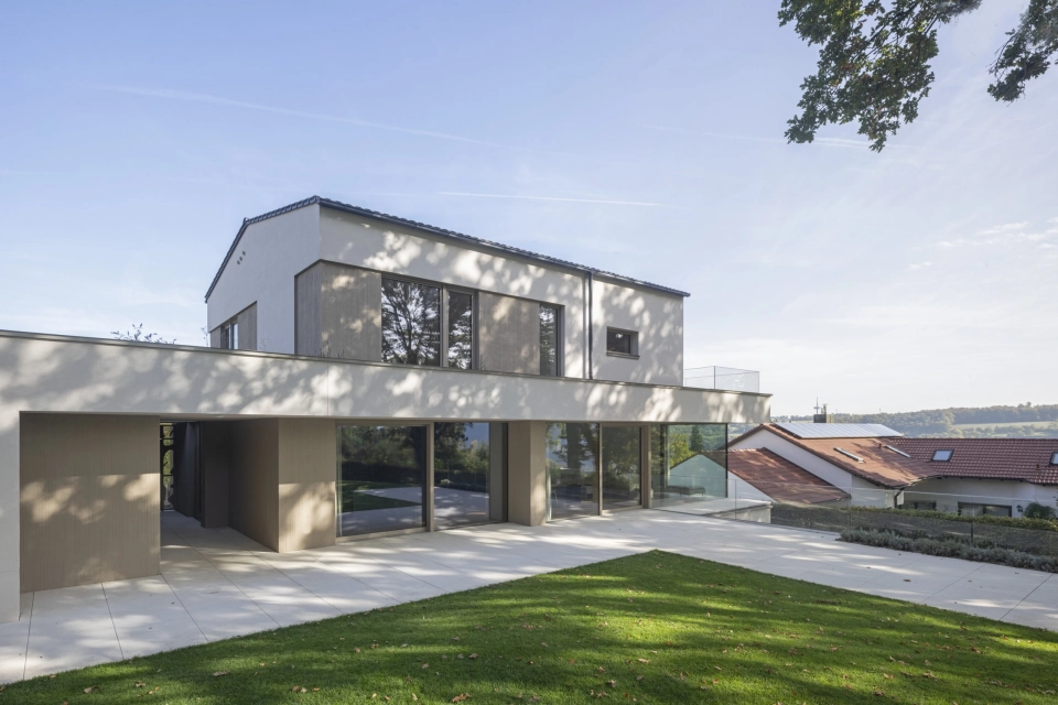 Rückseite eines hellen, modernen Wohnhauses, welches im Schatten hoher Eichen liegt. Im Vordergrund ist ein gepflegter Rasen und eine große Terrasse zu erkennen.