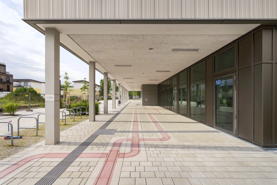 Überdachter Bereich eines großen Schulhofes gepflastert mit grau-beigen, versickerungsfähigen Pflastersteinen und einem roten Band, welches sich durch die Fläche zieht. Links ist ein begrünter Fahrrad-Parkplatz angelegt und rechts der Eingang zum Schulgebäude zu finden.