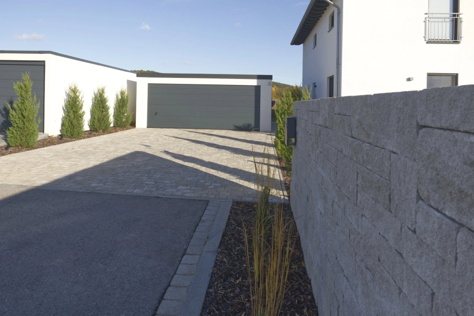 Einfahrt mit hellem GDM.VIA stone, welcher zu einer modernen Garage führt. Rechts verläuft eine GDM.MOLINA wall, daneben ein schmales Pflanzbeet mit Ziergräsern. Im Hintergrund stehen Garagen und eine Reihe immergrüner Gehölze entlang der Grundstücksgrenze.