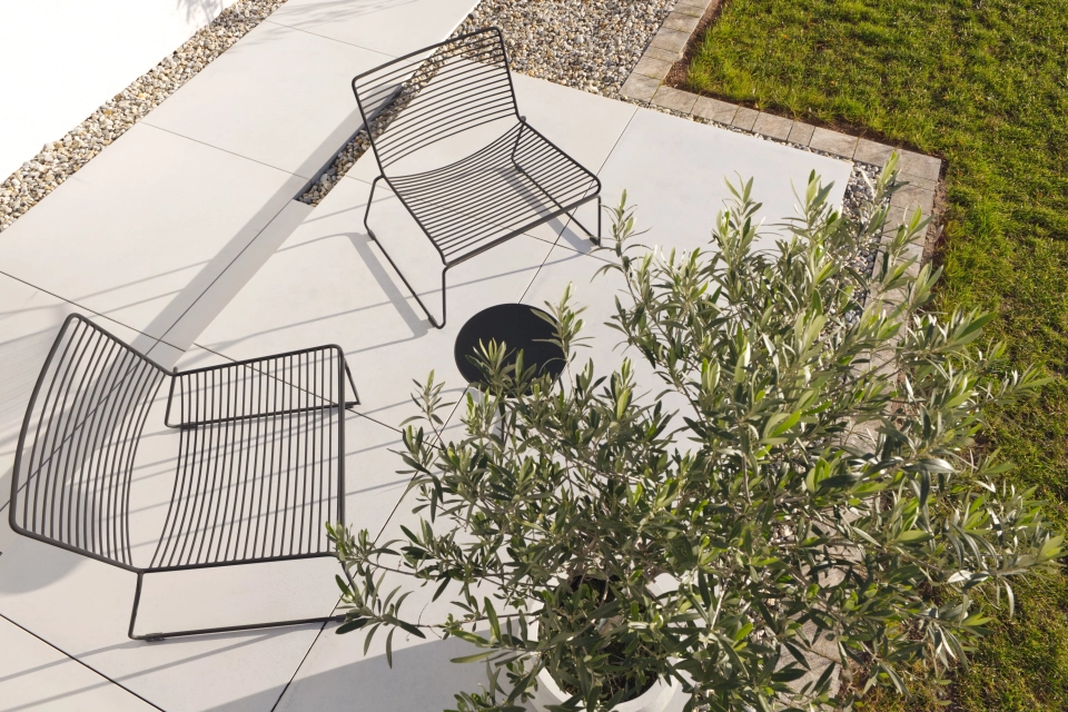 Terrassenbereich mit hellen GDM.MASSIMO light Terrassenplatten. Zwei filigrane Outdoor‑Stühle aus schwarzem Metall stehen auf der großzügigen Terrassenfläche. Im Vordergrund befindet sich ein Olivenbaum, dahinter sind ein Kiesstreifen sowie eine angrenzende Rasenfläche zu sehen.
