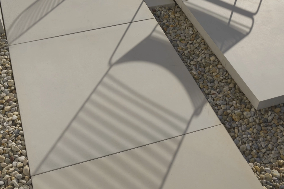Nahaufnahme der hellen GDM.MASSIMO light Platten, auf denen die Schatten eines filigranen Metallstuhls sichtbar sind. Die Platten grenzen an ein Kiesbett, das den Terrassenbereich umrahmt.