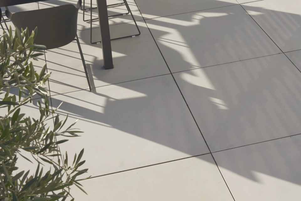 Modern gestaltete Terrasse mit hellen GDM.MASSIMO light Platten, auf denen ein dunkler Outdoor‑Esstisch mit Stühlen steht. Im Vordergrund ist ein bepflanztes Hochbeet in Corthenstahloptik zu sehen, im Hintergrund mehrere Kübelpflanzen und Schattenstrukturen auf der Plattenfläche.