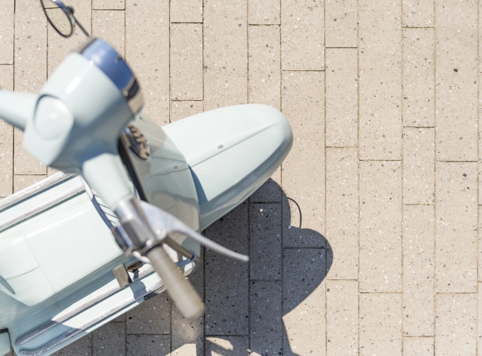 Draufischt auf eine himmelblaue Vespa auf einer geplasterten Fläche mit 
Terrazzo-Steinen mit sichtbarer Körnung Format eines Stabparkets.