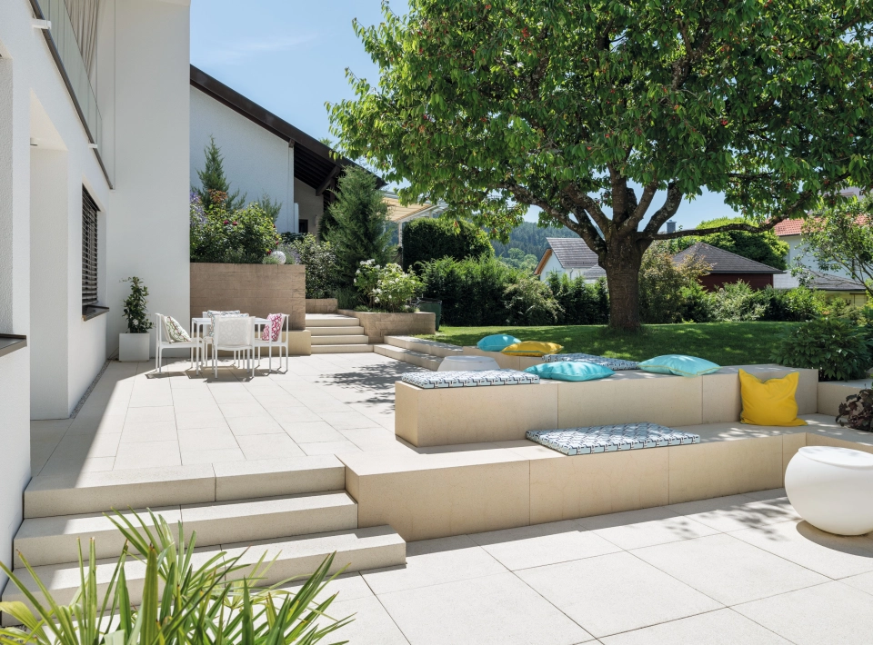Terrassenplatten in einem begrünten Garten mit einem großen Baum und Sitzelementen aus Beton.