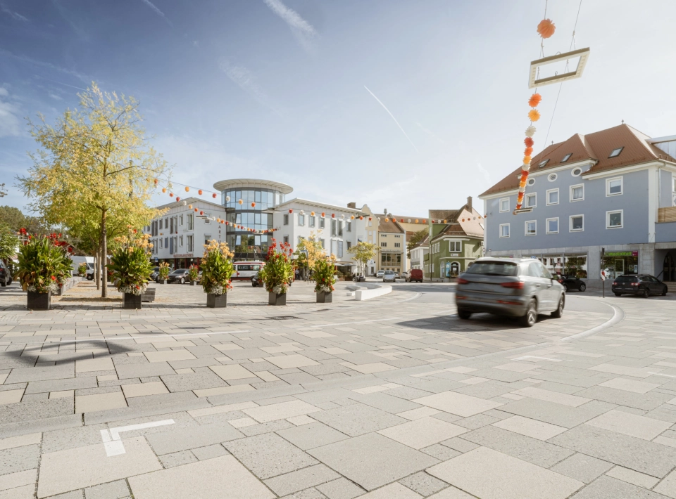 befahrbare Großfromatplatten im wilden Verband und abgestuften Grautönen auf einem Stadtplatz mit Bepflanzung.
